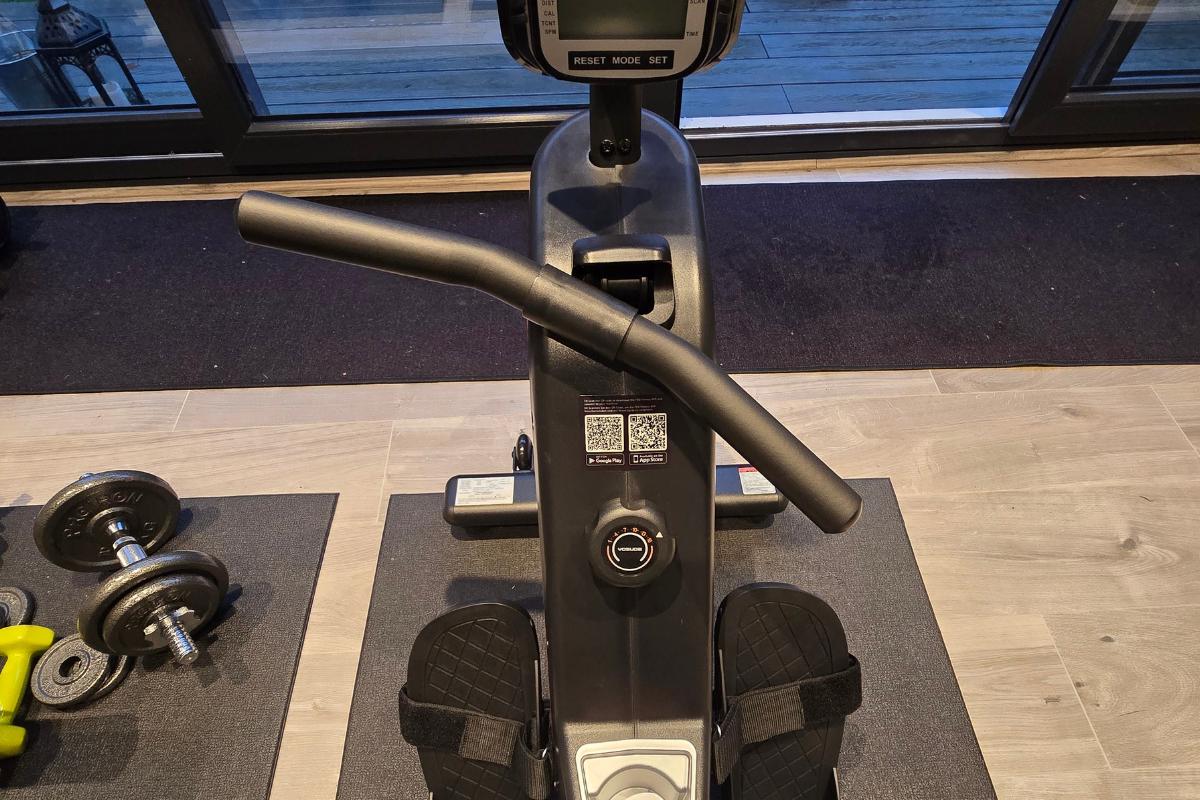 A view from above of a Yosuda RM4 rowing machine with foot pedals, a digital display, and handles. Dumbbells and gym mats are visible on the floor nearby, and a window is in the background—perfect for your next Yosuda RM4 review.