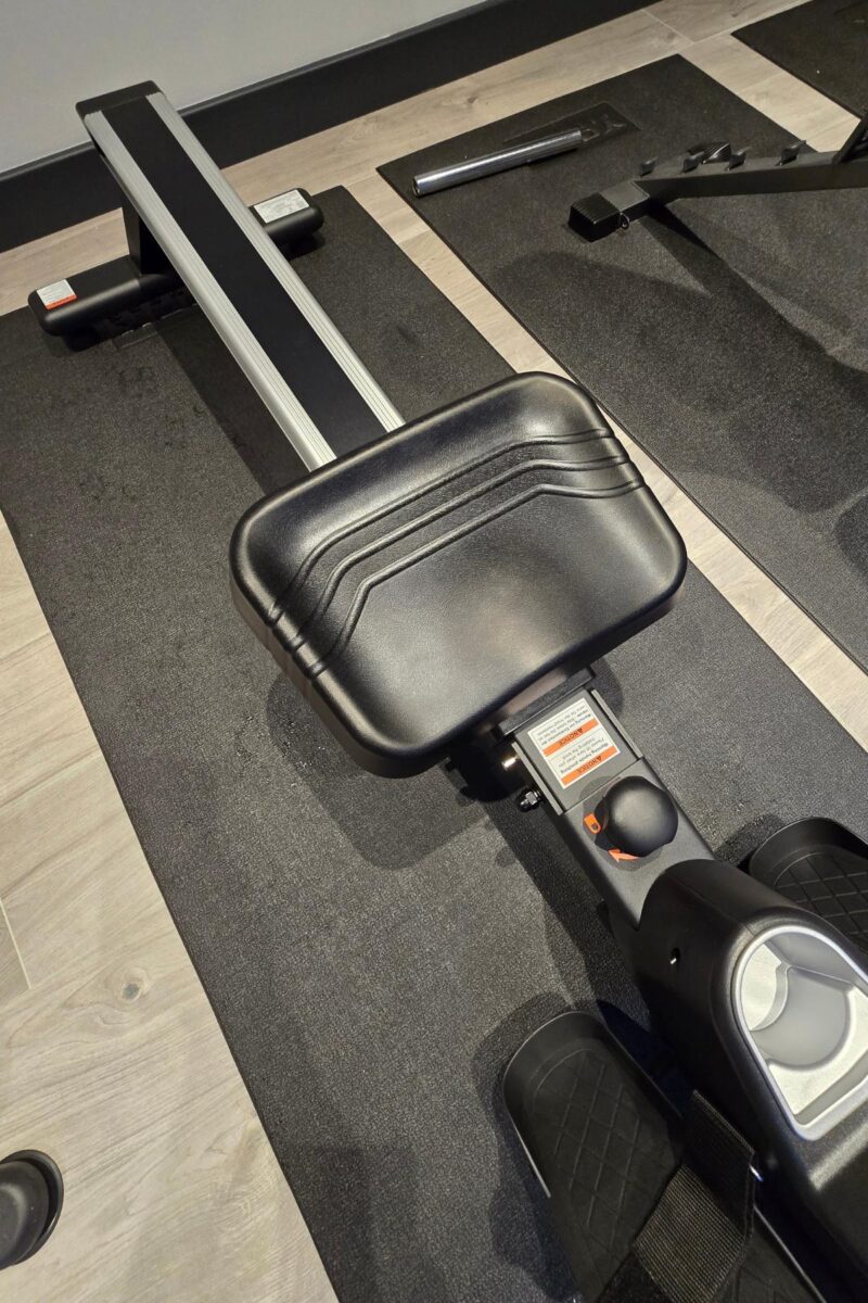 A close-up view of a Yosuda RM4 rowing machine seat and rail on a black exercise mat, positioned on a light wood floor in a gym setting—perfect for any Rowing Machine Guide.