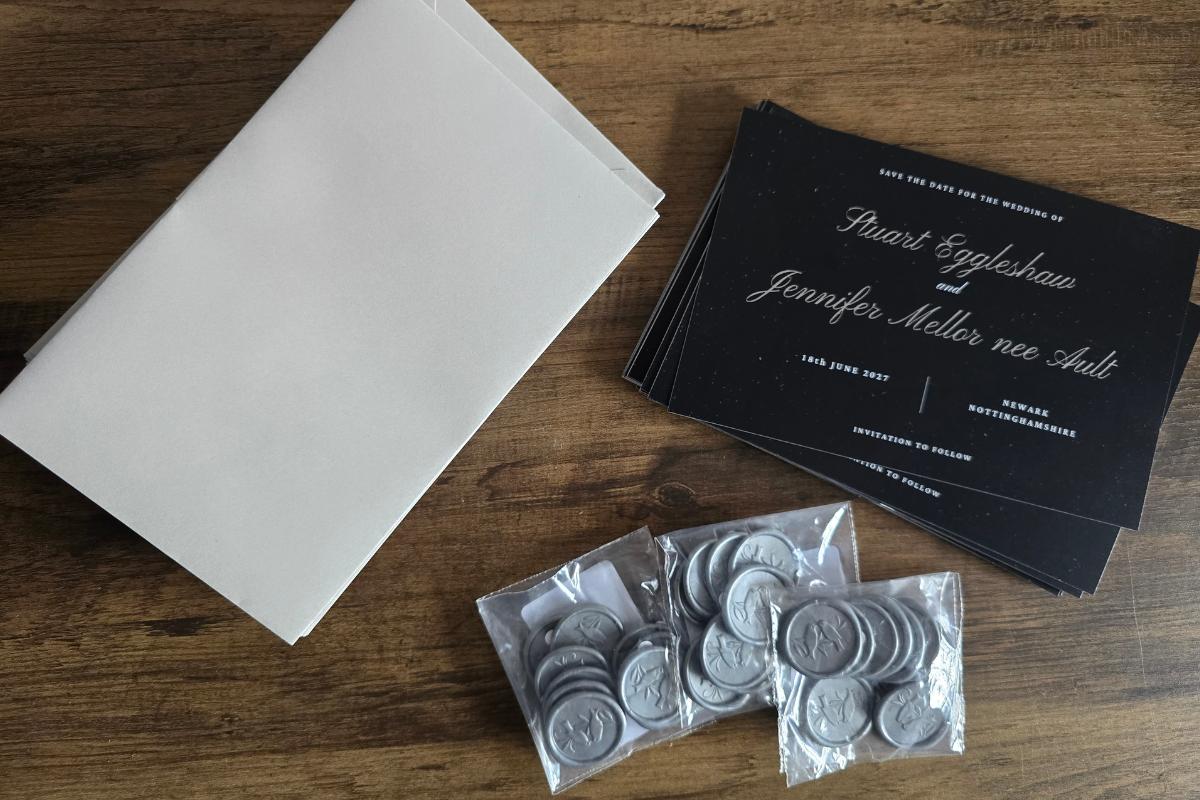 A stack of blank envelopes, several wedding invitations, and small packets of silver decorative coins are arranged on a wooden surface.