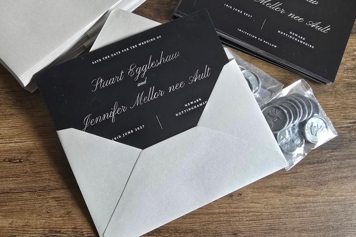 A black and white wedding invitation in an open envelope lies on a wooden surface, alongside save-the-date cards and silver wax seals stacked nearby. The wedding invitations feature elegant white text on a black background.