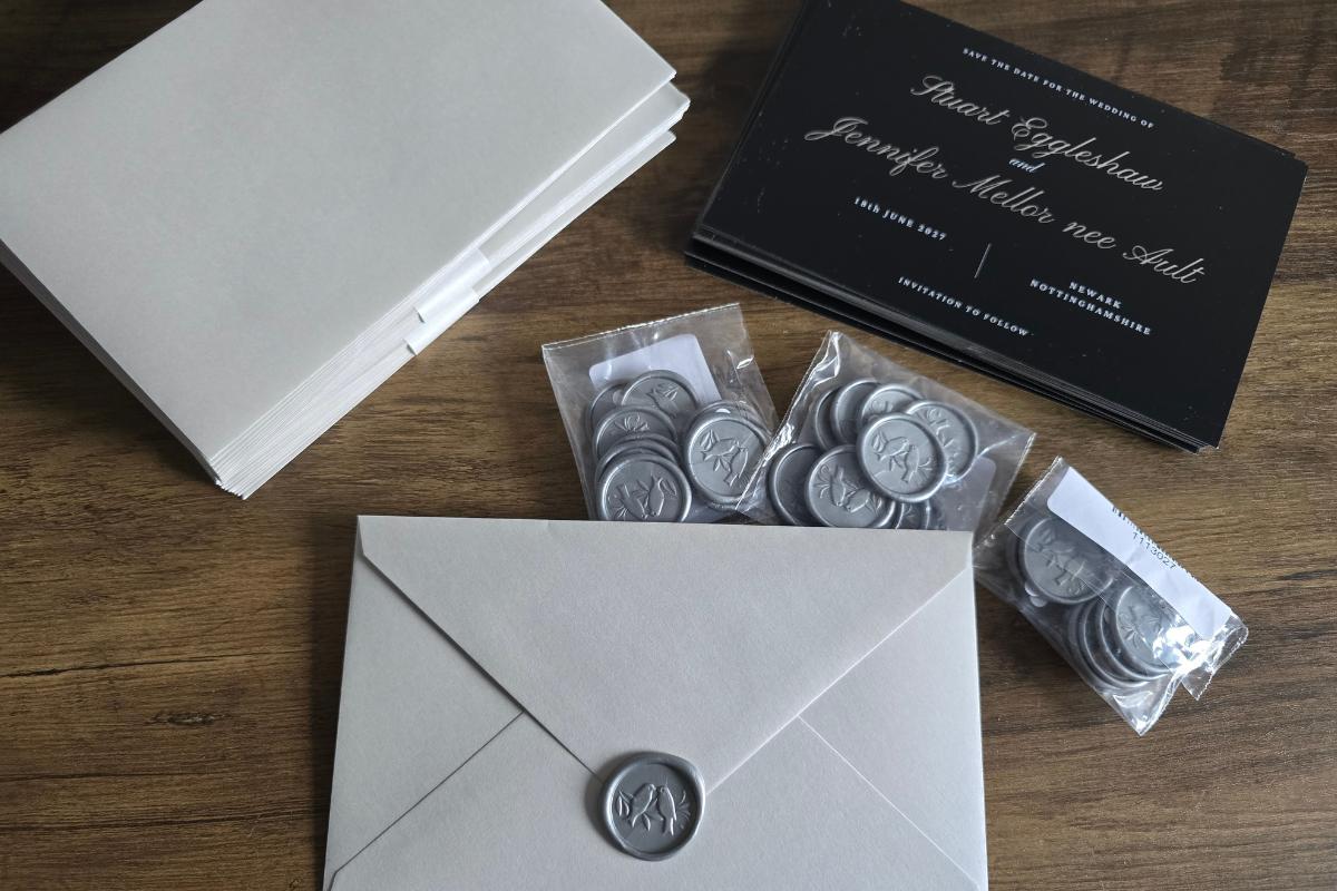 A stack of cream envelopes, one sealed with a grey wax seal, sits next to elegant wedding invitations and several packets of grey wax seals on a table. The black invitation features stylish white text perfect for your save-the-date cards.