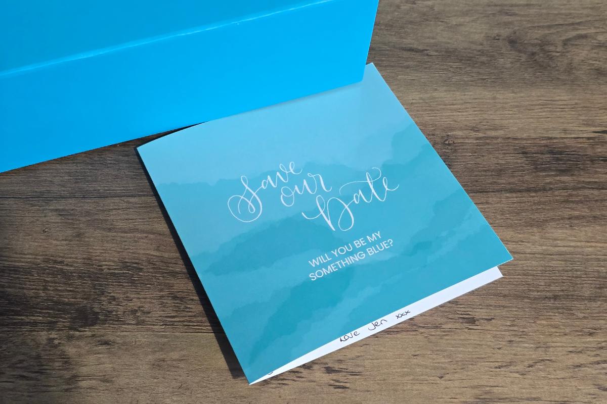 A blue "Save Our Date" card with white cursive text sits on a wooden surface next to a bright blue box. Perfect for capturing how I asked my bridesmaids, the card reads, "WILL YOU BE MY SOMETHING BLUE?" and is signed, "Love, LRA xxx.