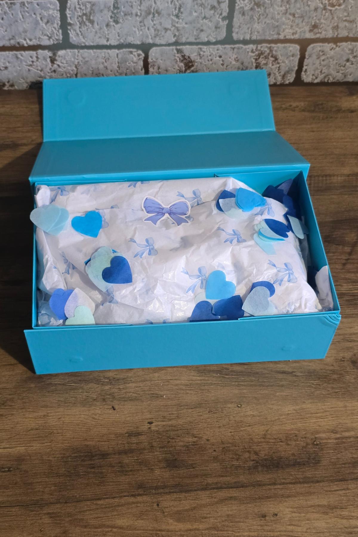 An open turquoise "Something Blue Ladies" box sits on a wooden surface. Inside, white tissue paper with blue and purple heart and butterfly cut-outs hints at how I asked my bridesmaids. The background features a light-coloured brick wall.