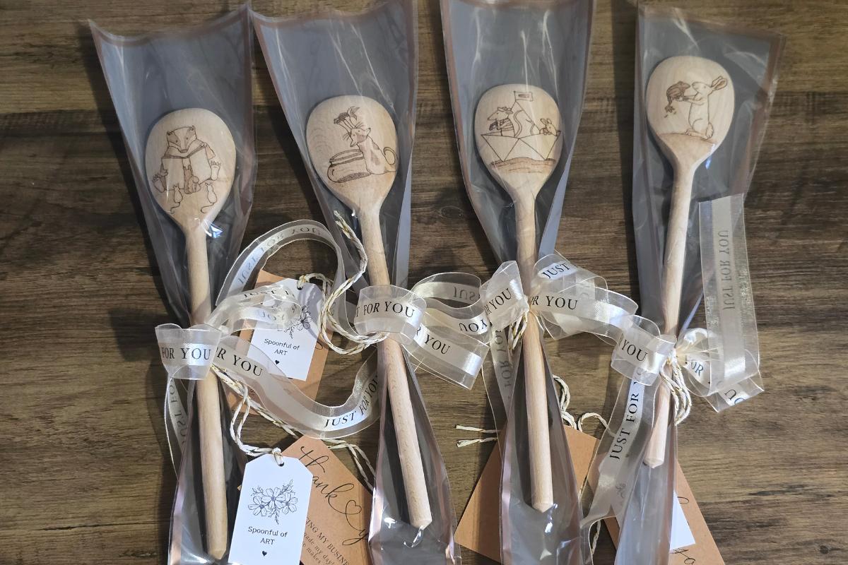 Five wooden spoons with engraved animal designs are individually wrapped in clear plastic, tied with ribbons and tags, and displayed on a wooden surface—perfect for those who love to shop small and support small businesses.