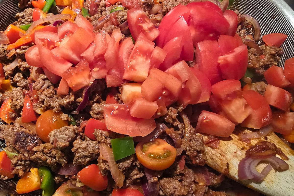 Minced beef is being cooked in a pan with chopped tomatoes, peppers, onions, and cherry tomatoes, all partially mixed together with a wooden spoon visible on the right—a healthy meal perfect to pair with spicy Cajun potato wedges.