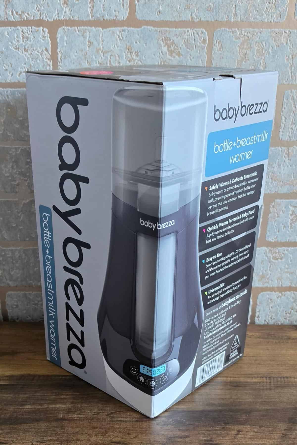 A box of the baby brezza bottle and breastmilk warmer sits on a wooden surface in front of a light brick wall. Ideal for parents seeking better sleep, the packaging highlights product features designed for babies in blue and white text.