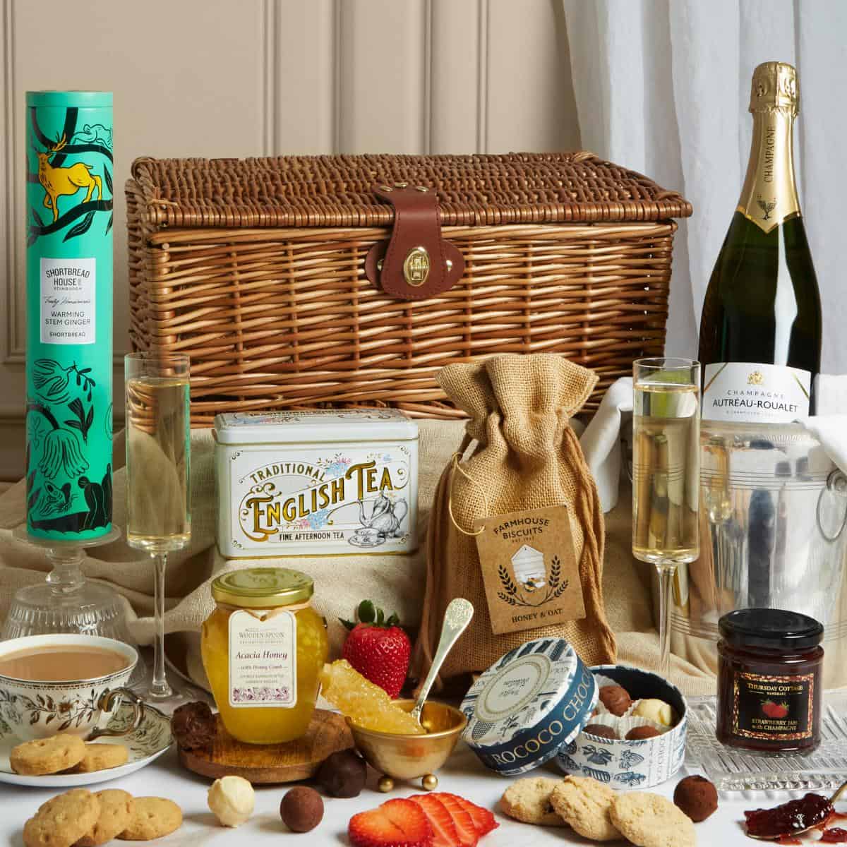 A wicker hamper perfect as an engagement gift or wedding present, featuring champagne in a bucket, two filled glasses, tea caddy, honey jar, biscuits, chocolates, preserves, cheese, teacup, strawberries, and assorted treats on a table.