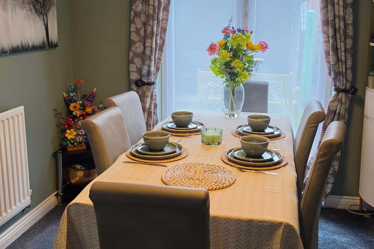 A dining table set for four with grey chairs, green crockery, and a beige tablecloth awaits a family easter celebration. A glass vase with colourful flowers is in the centre as sunlight streams through sheer curtains in the background.