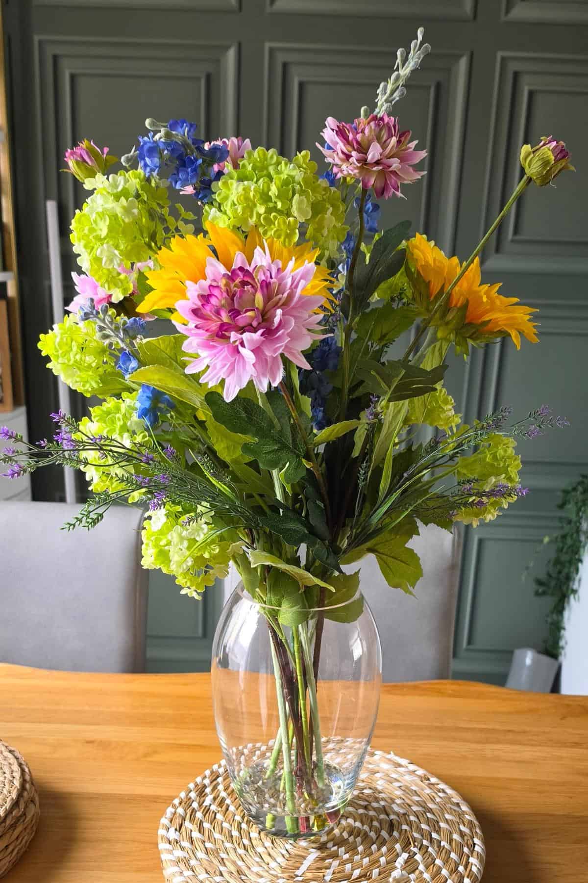 A clear glass vase holds a vibrant sunflower mix of pink dahlias, yellow floralsilk faux sunflowers, blue and green blooms, and greenery, set on a woven mat atop a wooden table with a green panelled background.