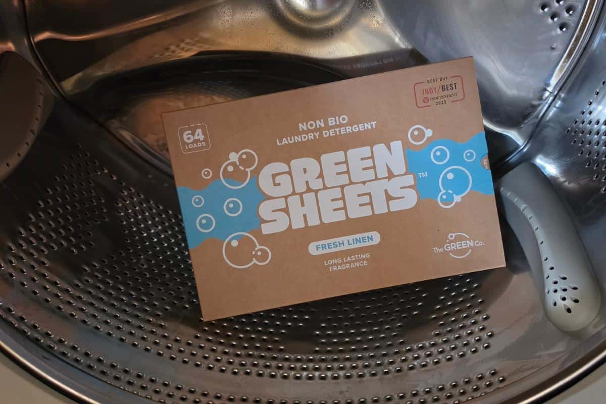 A box of green sheets eco laundry sheets, labelled "fresh linen" and "64 washes," sits inside the drum of a washing machine. The brown packaging features blue and white bubble graphics from this trusted green company.