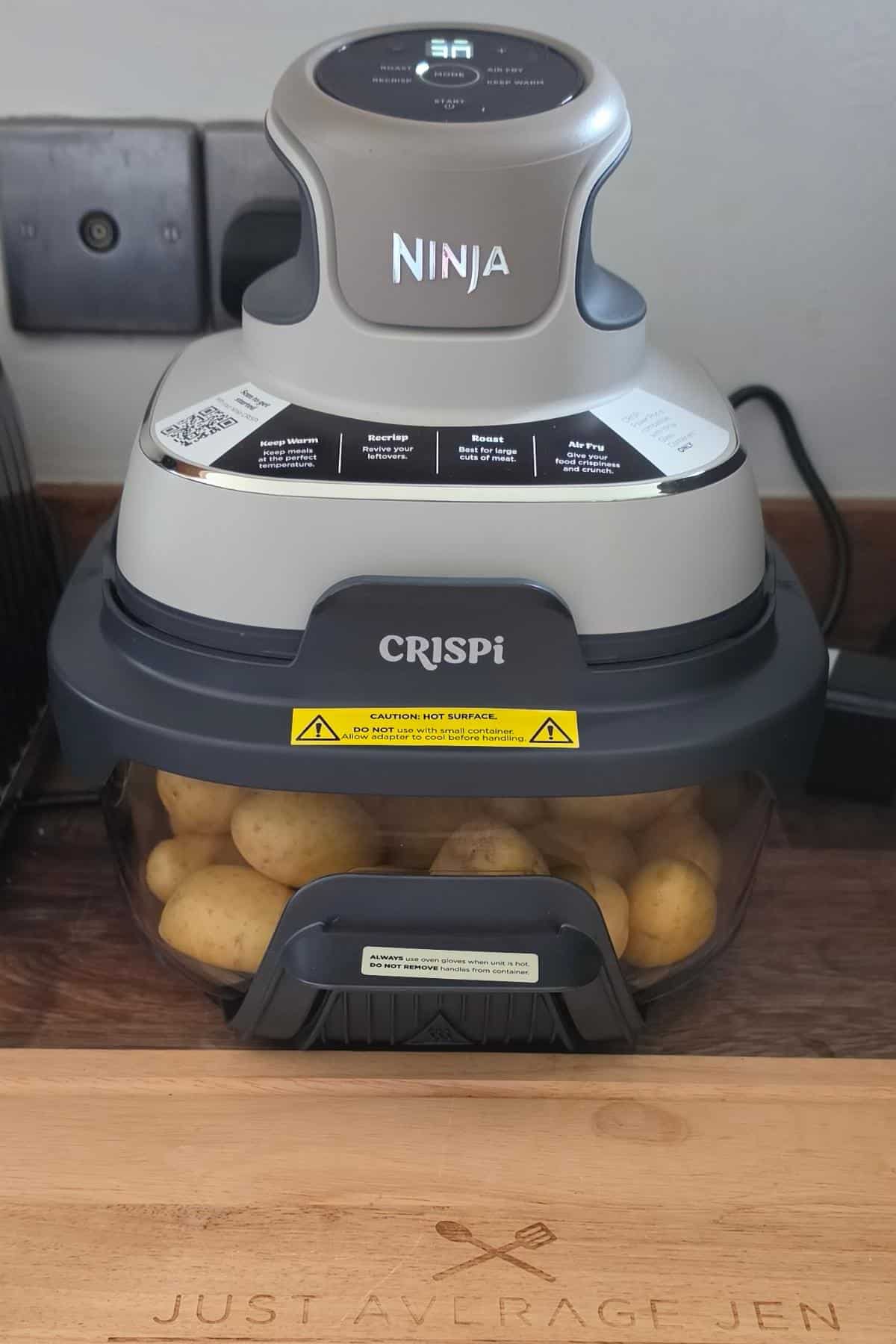 A ninja air fryer labelled "crispi" sits on a kitchen counter, ready for an amazing easter feast with whole, uncooked potatoes inside. The appliance is plugged in and resting on a wooden board engraved with "just a large jen.