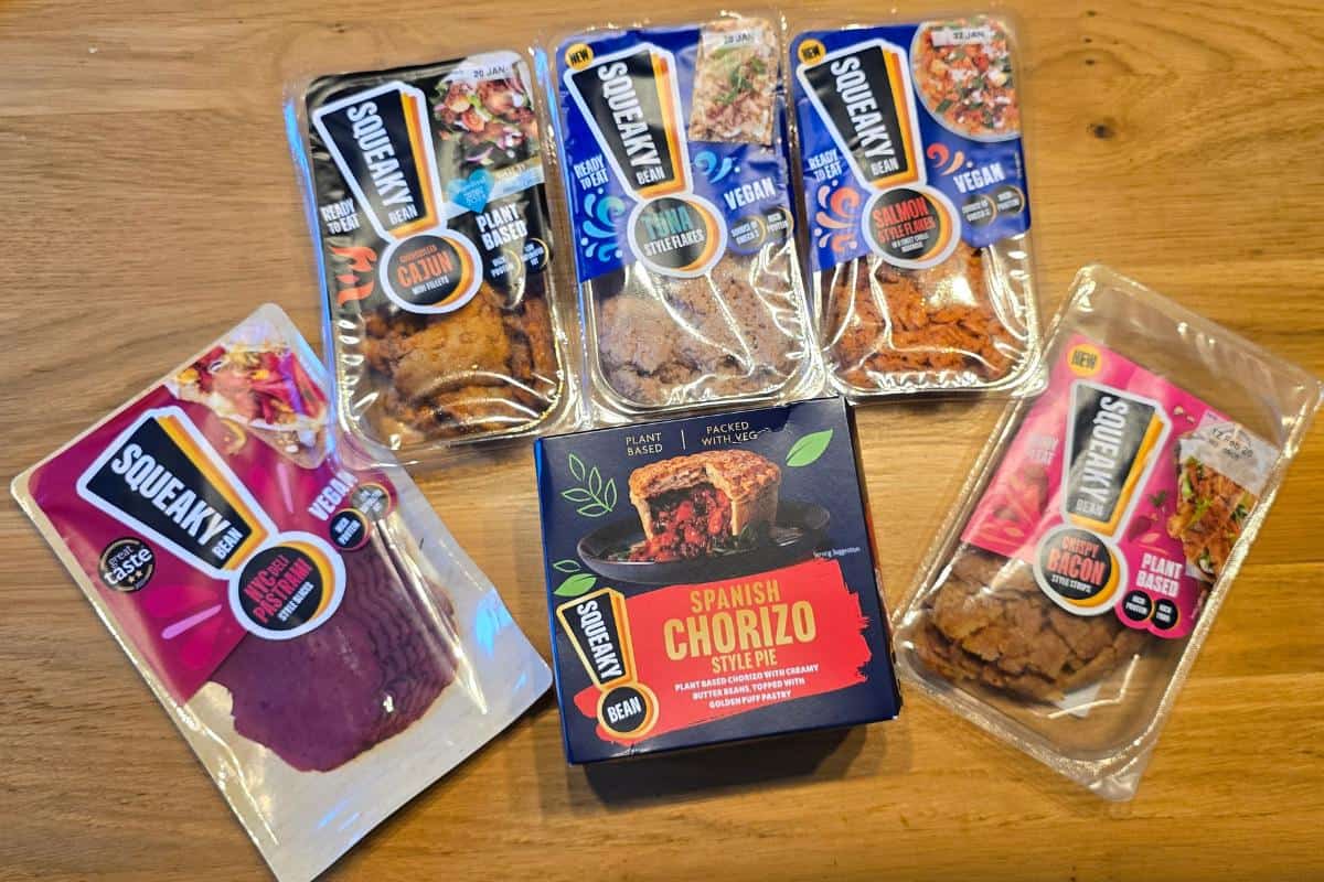 Five packs of squeaky bean plant-based meat products, perfect for vegan swaps, are arranged on a wooden surface. The packaging features colourful labels with enticing images of the food.