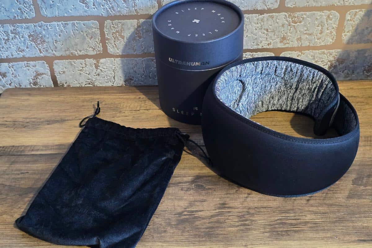 A black ultrahuman sleep mask with a padded interior is displayed on a wooden surface next to a black drawstring pouch and the cylindrical product box. Perfect for restful nights—stay tuned for our review and an exciting giveaway! A brick wall is in the background.