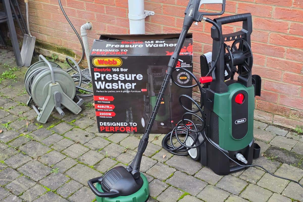A green and black webb wepw2200 electric pressure washer stands on a patio near its box. Hoses and cables are attached, with a hose reel nearby against a brick wall—ideal for decking cleaning or patio cleaning.