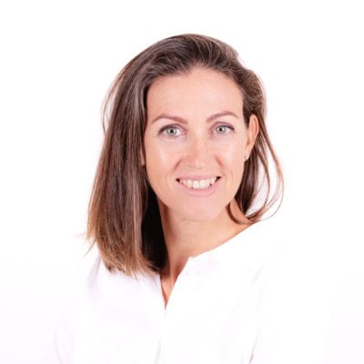 A woman with straight, shoulder-length brown hair smiles at the camera. She is wearing a white shirt and, embracing her perimenopause journey, is posed in front of a plain white background.