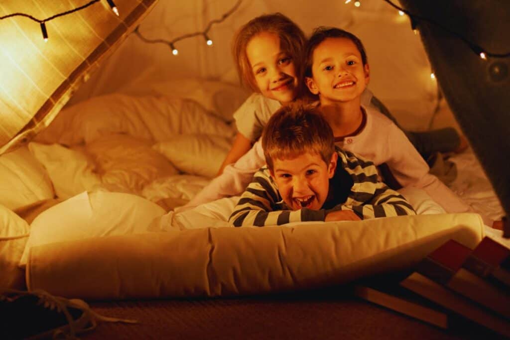 Three smiling children are lying on a bed inside a cosy blanket fort, surrounded by soft pillows and warm fairy lights. This playful scene is perfect for rainy day fun and creative indoor activities for children, with books stacked nearby.