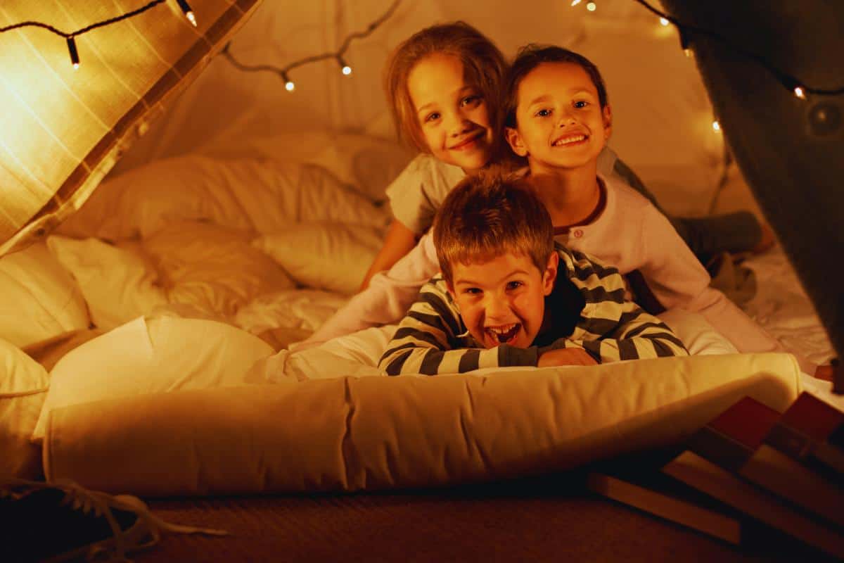 Three smiling children are lying on a bed inside a cosy blanket fort, surrounded by soft pillows and warm fairy lights. This playful scene is perfect for rainy day fun and creative indoor activities for children, with books stacked nearby.