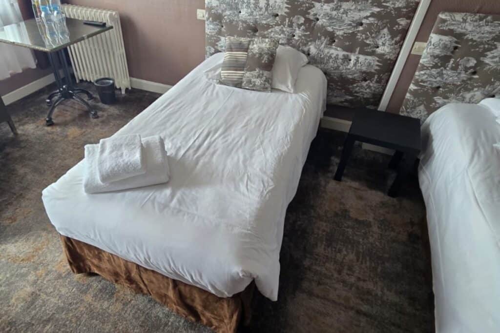 A neatly made single bed with white linen, two pillows, and folded towels on top. The bed is next to a small black bedside table and another bed in a carpeted room with patterned wallpaper, creating a cosy retreat in the heart of montreuil-sur-mer.