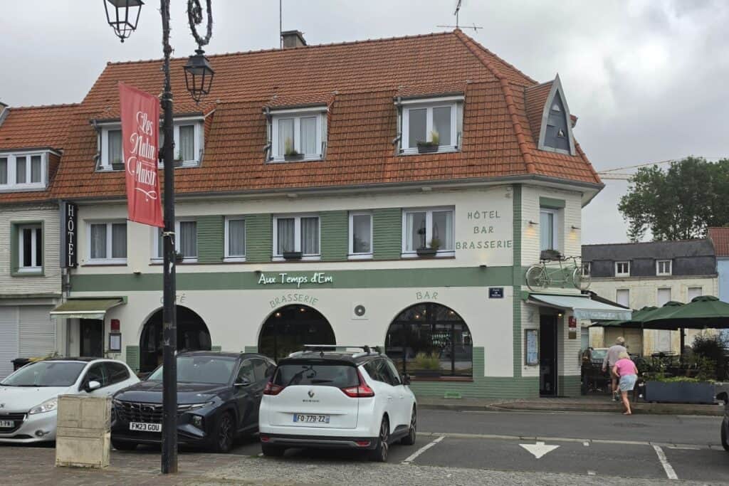 A street view of a french brasserie in montreuil-sur-mer with a red-roofed building, parked cars, a hotel sign, and people walking near outdoor seating under green umbrellas. The sky is overcast.
