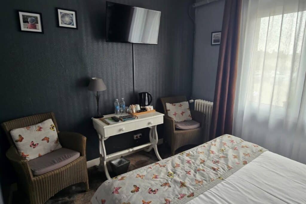 A cosy hotel room in montreuil-sur-mer with two wicker chairs, a small desk with water bottles, a kettle, and a lamp, a bed with butterfly bedding, and a tv mounted on a dark wall beside a window with sheer curtains.