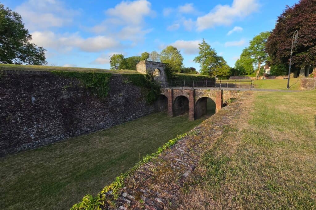 A brick bridge crosses a grassy moat next to a tall stone wall in montreuil-sur-mer, surrounded by green trees and a blue sky with clouds.