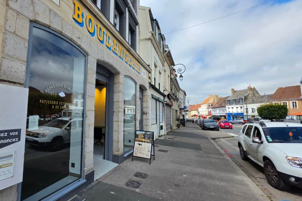A street view of a french bakery with "boulangerie" in yellow letters, open door, parked cars, and neighbouring shops on a bright day in the small town of montreuil-sur-mer.