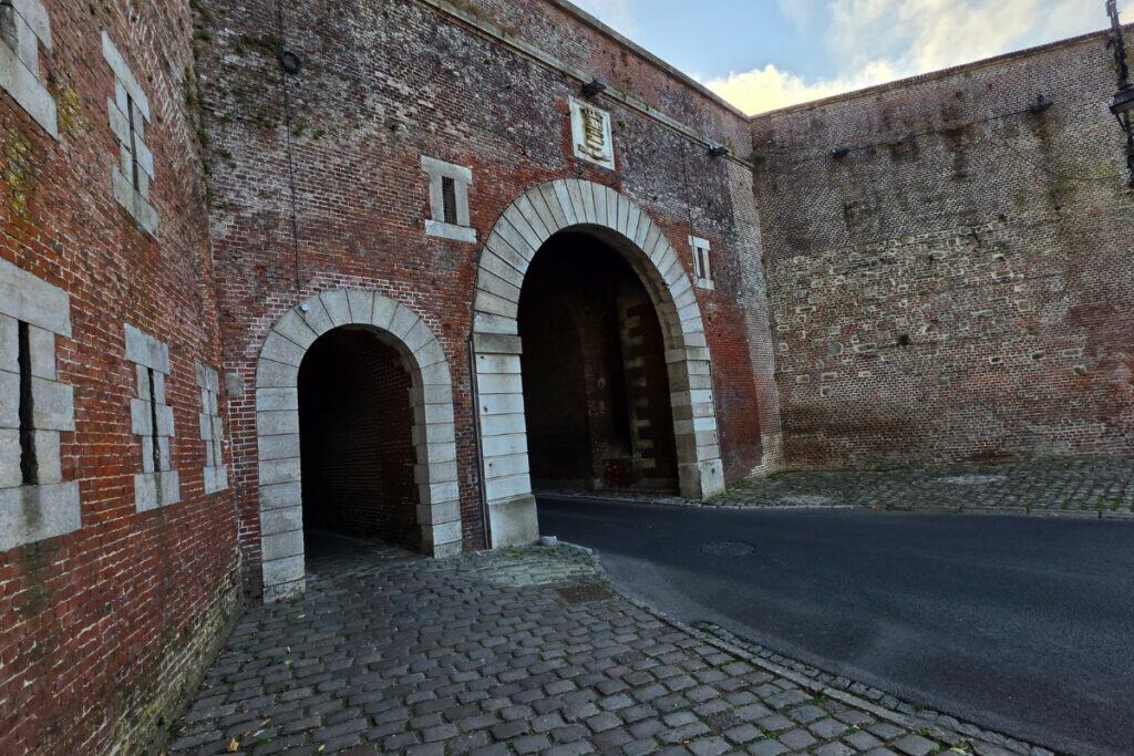 Two arched doorways in a tall brick wall, one large and one small, stand beside a paved road and cobblestone pavement in montreuil-sur-mer. The wall shows signs of age, with moss and weathering visible.