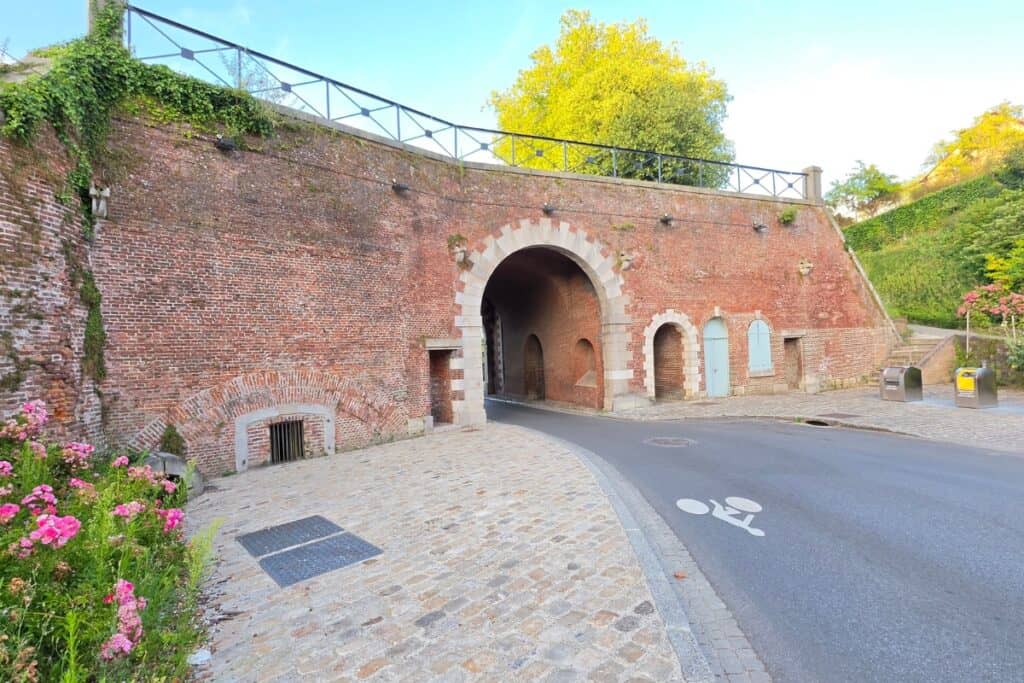 A cobblestone road curves through a large brick archway in an old city wall in montreuil-sur-mer, surrounded by greenery and flowers, with cycle lane markings on the road.