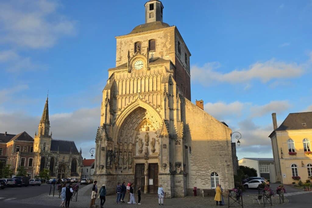 A group of people gather in front of a tall, stone church with a clock tower and ornate entrance in montreuil-sur-mer, under a blue sky with some clouds. Other historic buildings and a spired church are visible in the background.