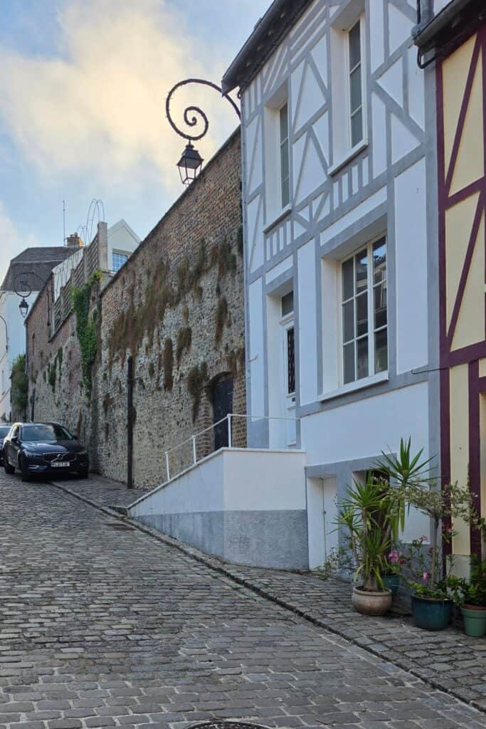 Cobblestone street with a parked car, half-timbered houses, potted plants, and an old brick wall covered in ivy—a charming scene reminiscent of montreuil-sur-mer. A vintage street lamp hangs above, and the sky is partly cloudy.