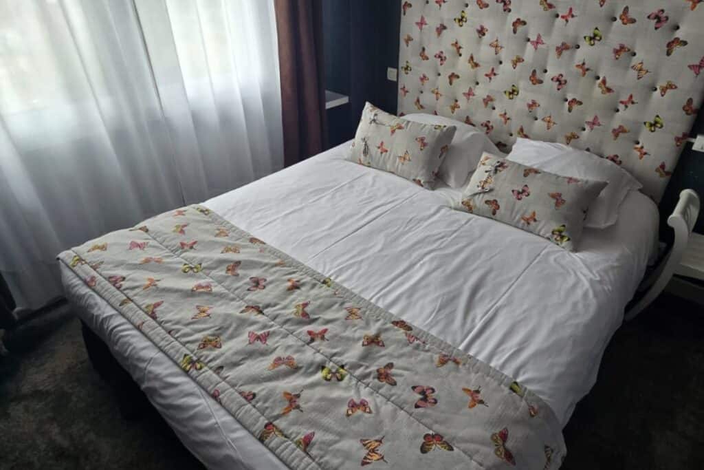 A neatly made bed with white sheets and butterfly-themed pillows, headboard, and blanket brings a touch of charm to this inviting bedroom. Set by a window with sheer white curtains that let in natural light, the room evokes the tranquil ambience of a cosy retreat in montreuil-sur-mer.
