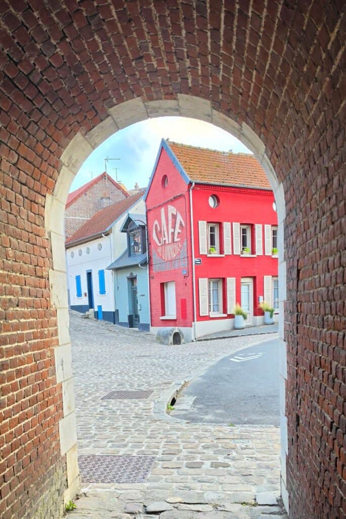 View through a brick archway to a cobbled street in montreuil-sur-mer, featuring a vibrant red building with “safe universe” painted on its side. Other colourful houses and clear sky are visible in the background.