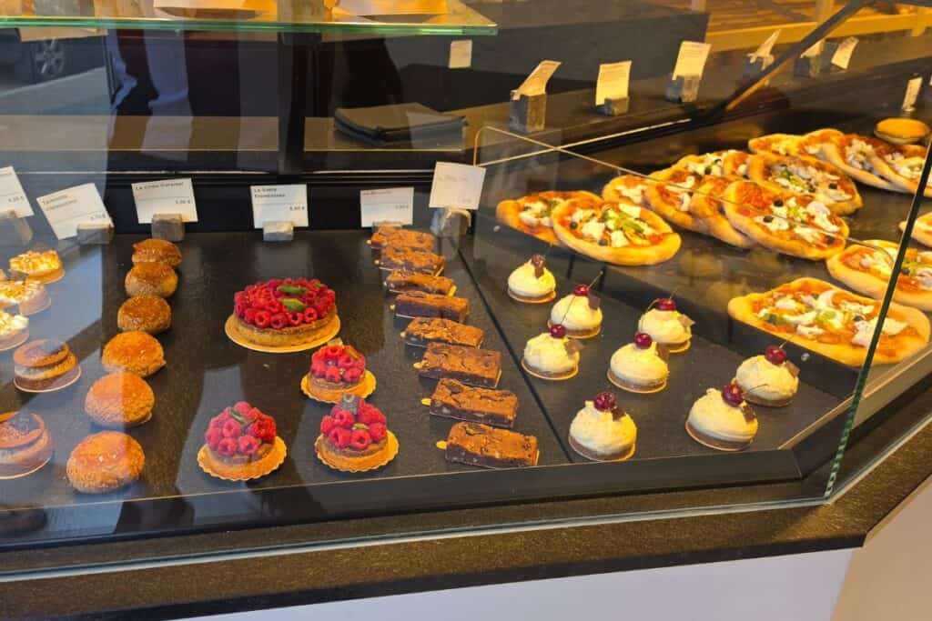 A display case with assorted pastries, including fruit tarts, chocolate desserts, cream puffs, and mini pizzas—all neatly arranged with price tags behind the glass—evokes the charm of a montreuil-sur-mer bakery.