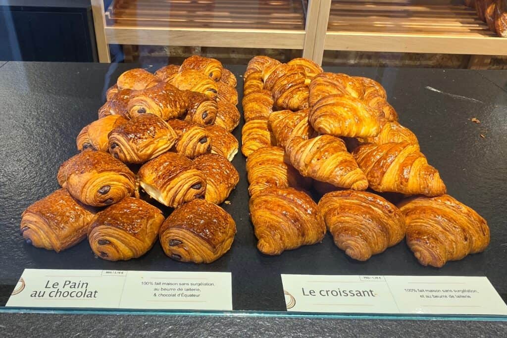 Rows of golden-brown pain au chocolat and croissants are displayed on a bakery counter in montreuil-sur-mer, with labels in french in front of each row.