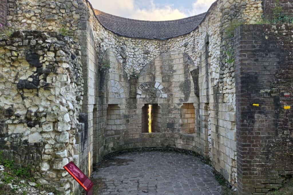 Old stone fort wall with an arched design, two narrow windows, and weathered bricks in Montreuil-sur-Mer. A small red information sign stands on the left amid rough stones. The ground is paved with uneven stones.