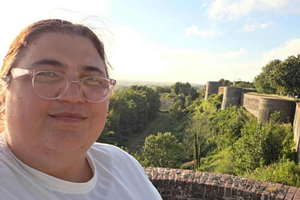 A person wearing glasses and a white shirt smiles at the camera with the ancient stone fortress walls of montreuil-sur-mer, lush greenery, and a bright sky in the background.