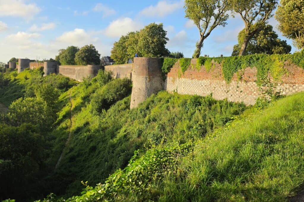 A historic stone and brick fortification wall stretches along a grassy, overgrown embankment in montreuil-sur-mer, with trees and greenery surrounding it under a partly cloudy blue sky.