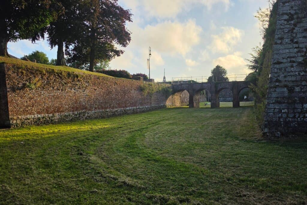 Grassy moat bordered by old stone and brick fortress walls, with an arched stone bridge in the background, evokes the historic charm of montreuil-sur-mer. Trees and a lamp-post are visible above the walls under a partly cloudy sky.