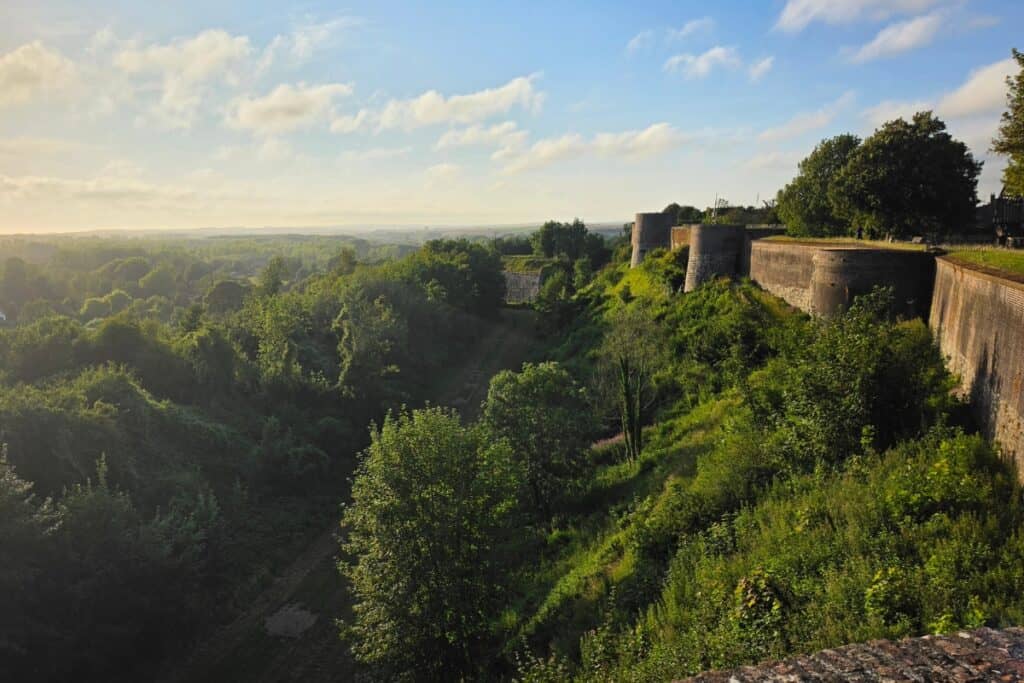 Old stone fortress walls border a lush, green hillside under a blue sky with scattered clouds in montreuil-sur-mer. Trees and dense vegetation fill the landscape, and the sunlight casts a warm golden glow across the scene.