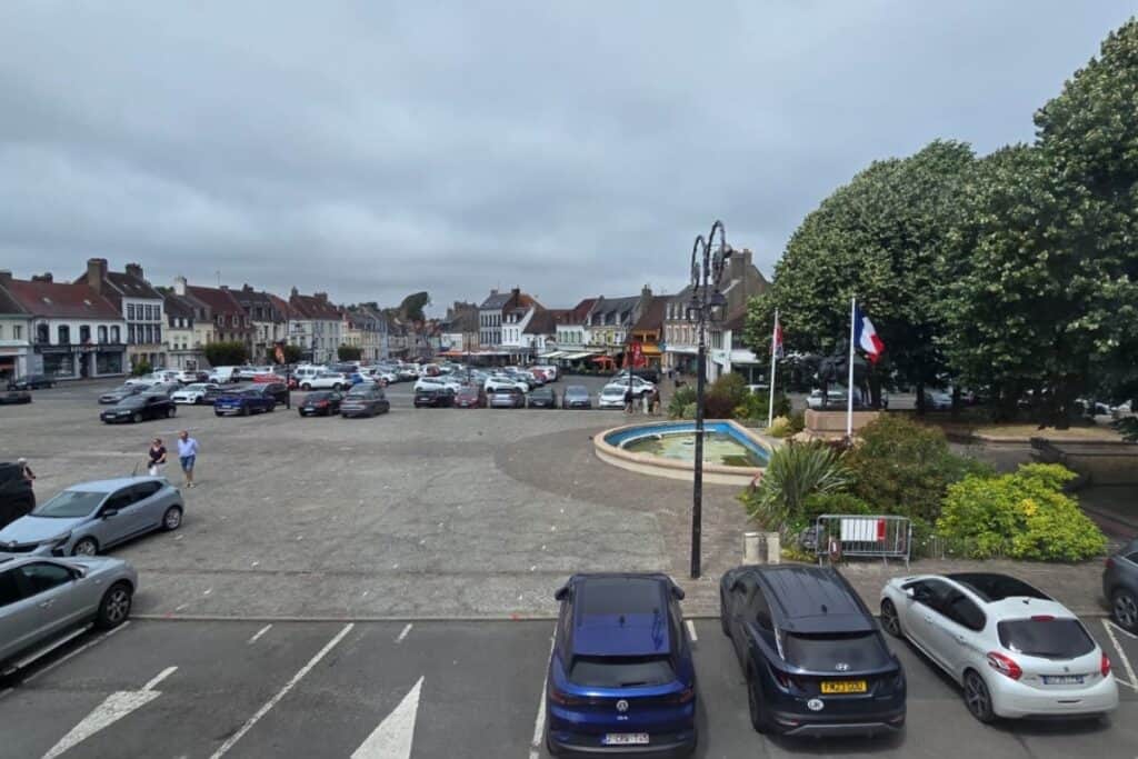A town square in montreuil-sur-mer with parked cars, a fountain, trees, the french flag, and surrounding buildings under a cloudy sky.
