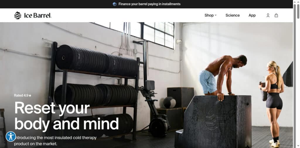 A man climbs onto a large black barrel in a gym whilst a woman in sportswear watches, both surrounded by gym equipment and natural light from tall windows.