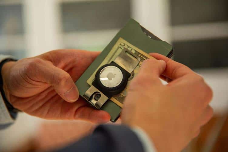 A person holds a green wallet with cash, an apple airtag, and a metal clip attached, focusing on the hands and wallet in a home setting—showcasing how even traditional wallets can incorporate modern features alongside the sturdiness often seen in metal wallets.