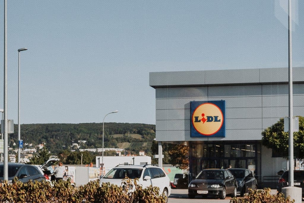 A lidl supermarket with cars parked in front, set against a backdrop of green hills and clear blue sky.