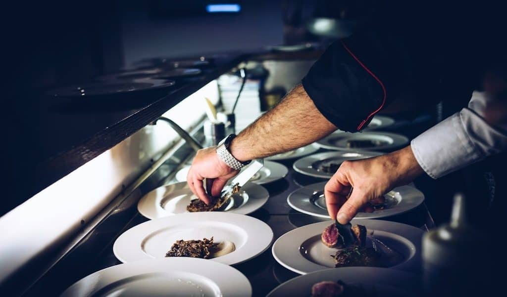 A chef is plating food onto several white dishes lined up on a counter under bright lighting.
