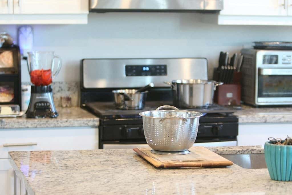 A stainless steel colander on a wooden chopping board sits on a kitchen worktop; saucepans are on the hob and a blender with red contents is nearby.