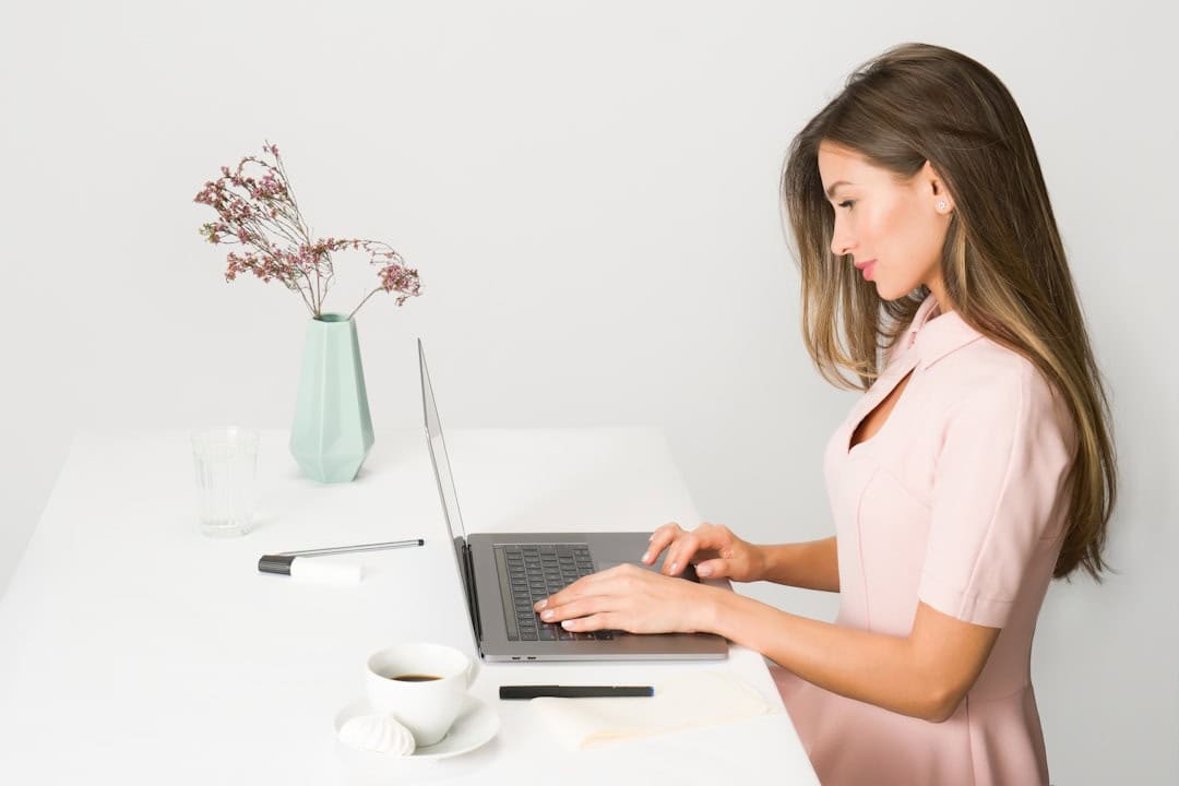 Woman in a pink dress types on a laptop at a white desk with a cup of coffee, pen, and a vase with dried flowers.