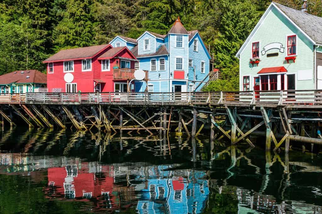 Colourful wooden houses on stilts stand by the water at ketchikan, reflecting in the calm surface, with forested trees in the background.