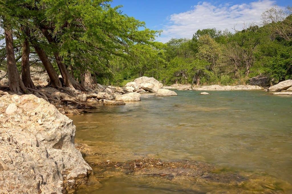 Clear river with shallow water flows past large rocks and leafy trees under a partly cloudy blue sky in a natural outdoor setting.
