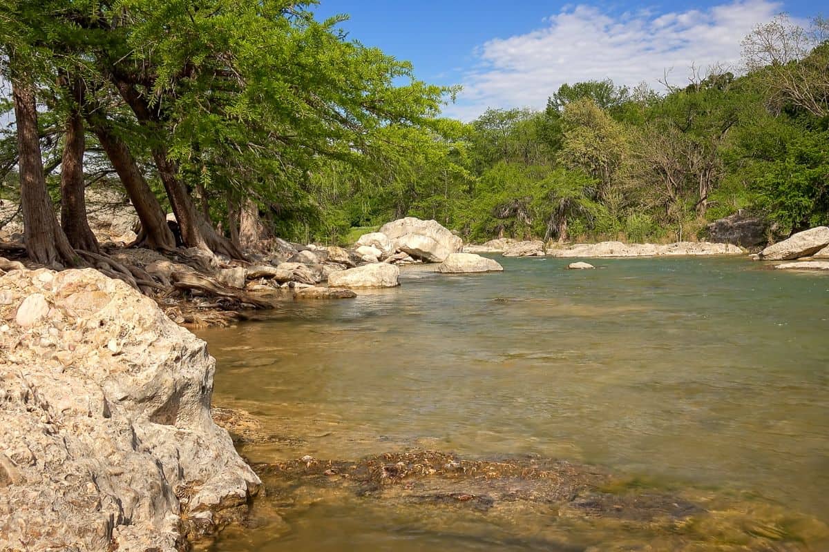 Clear river with shallow water flows past large rocks and leafy trees under a partly cloudy blue sky in a natural outdoor setting.