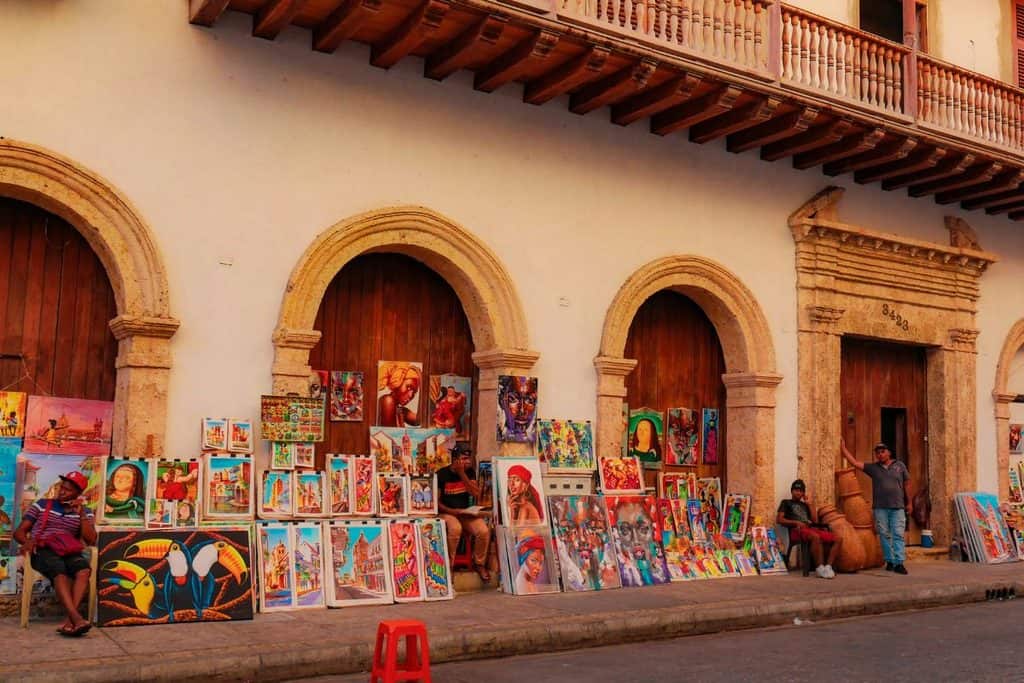 Street artists display colourful paintings for sale outside a historic building with arches and wooden doors; a few people sit and stand nearby.